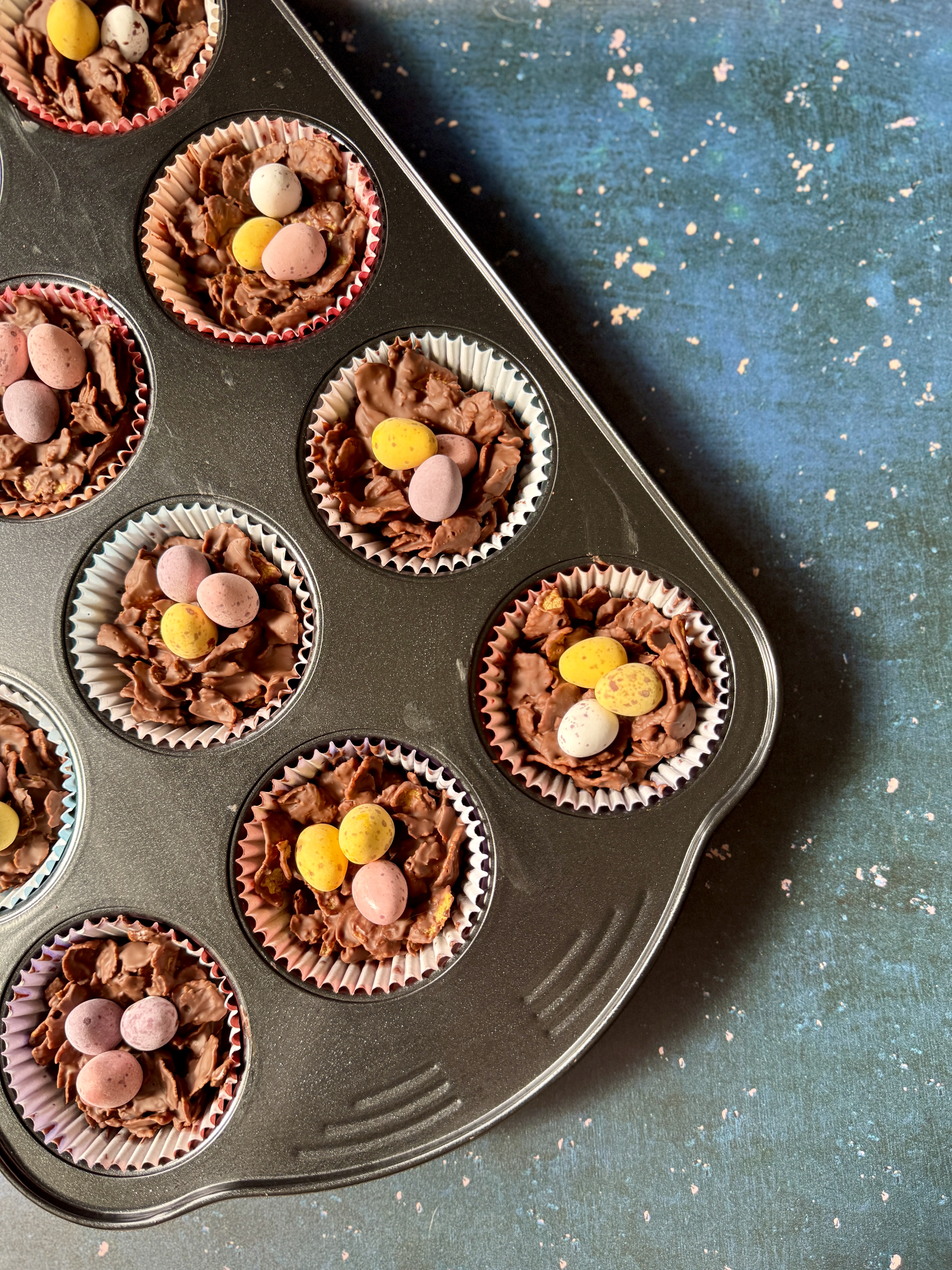 Homemade Easter cornflake cakes in pastel paper cases, arranged in a cupcake tray on a blue textured background.